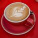 A beautifully crafted cappuccino with intricate latte art served on a vibrant red table.